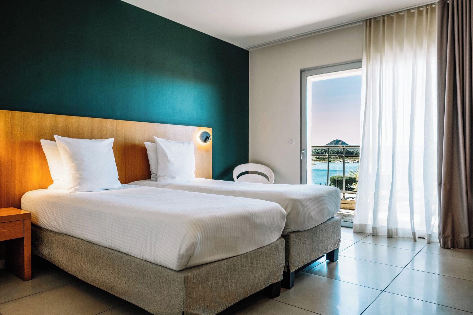 A hotel room with two beds, a nightstand, a chair, and a balcony with a view of the sea.