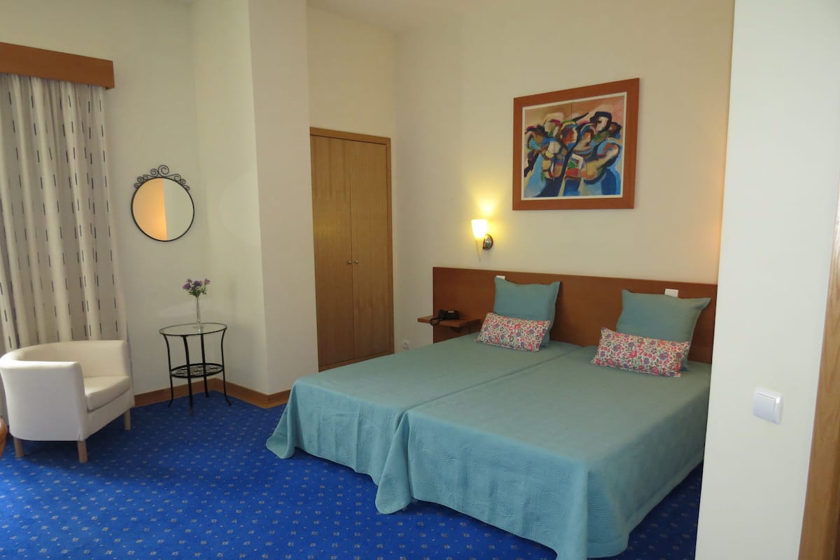 A hotel room with a bed, a chair, a small table with a vase, a door, and a painting on the wall.