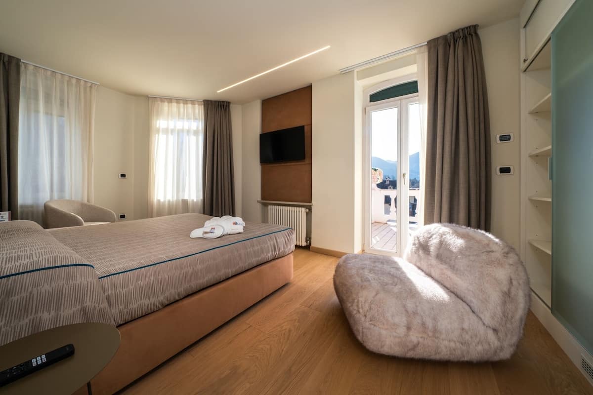 A hotel room with a bed, a chair, a fluffy ottoman, a TV, and a view of the outdoors.