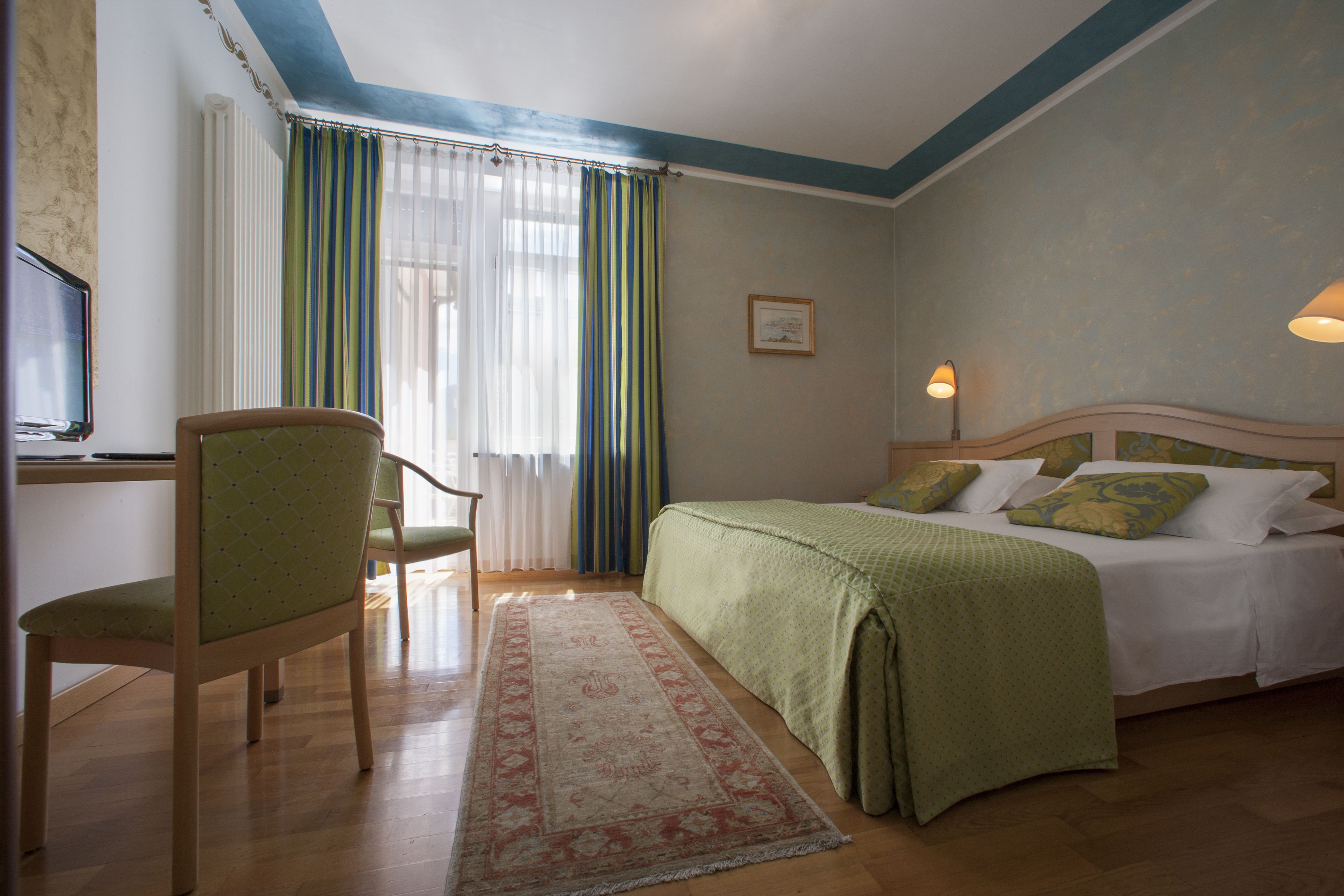 Ein Hotelzimmer mit Bett, einem Sessel, einem Schreibtisch mit Computer und einem Fenster mit Vorhängen.