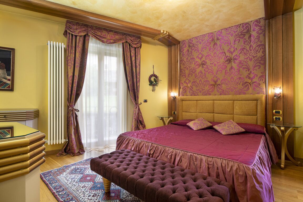 Quarto com cama grande, cabeceira de madeira, um banquinho, radiador, cortinas e parede estampada.