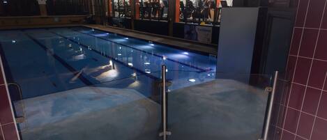 A large indoor swimming pool with a hot tub area, surrounded by a tiled wall and a glass barrier.