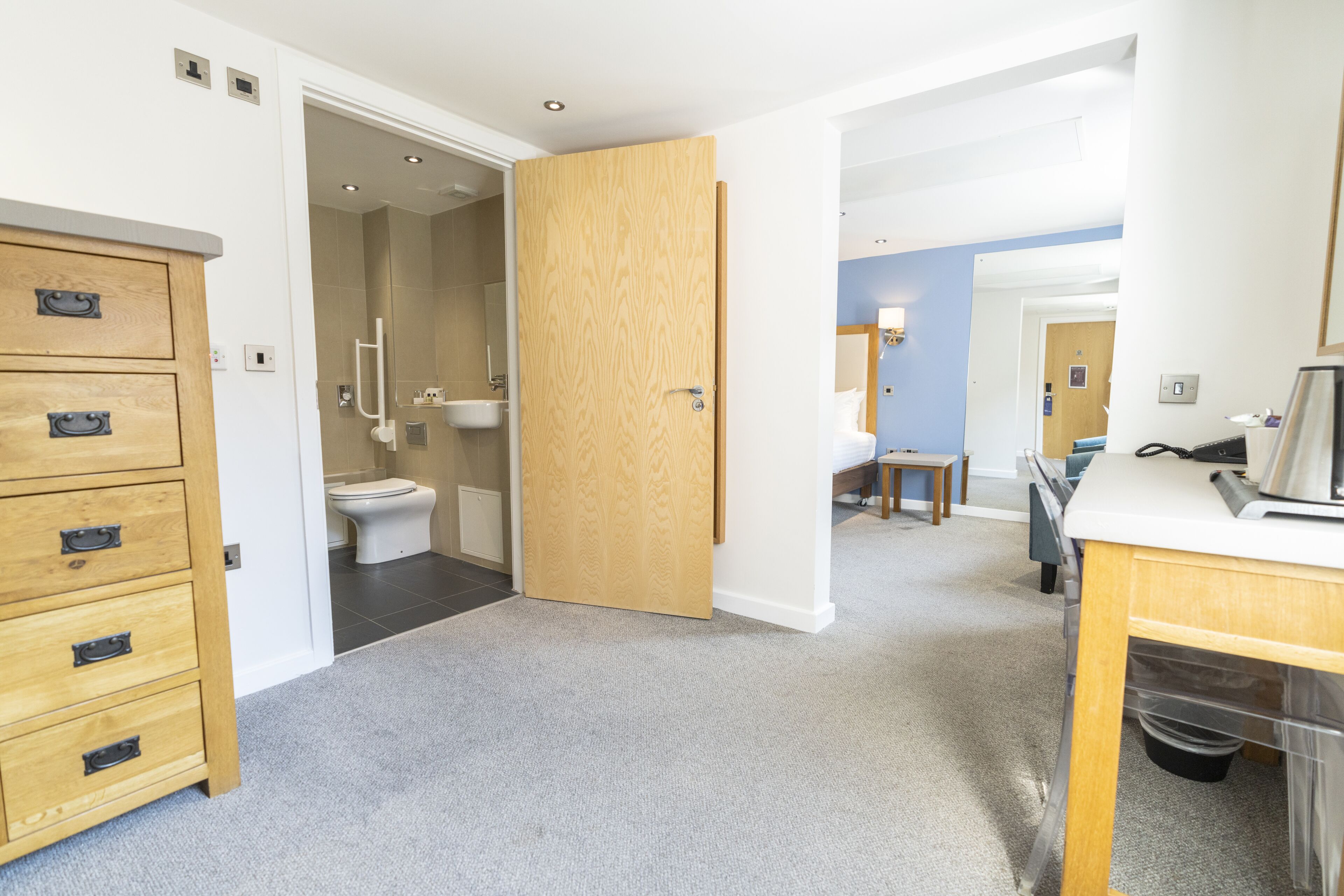 A hotel room with a wooden dresser, a bathroom with a toilet and sink, and a small table with a chair.
