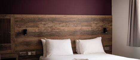 Una cama bien tendida con sábanas blancas y una toalla doblada, cabecera de madera y panel de interruptores en la pared.