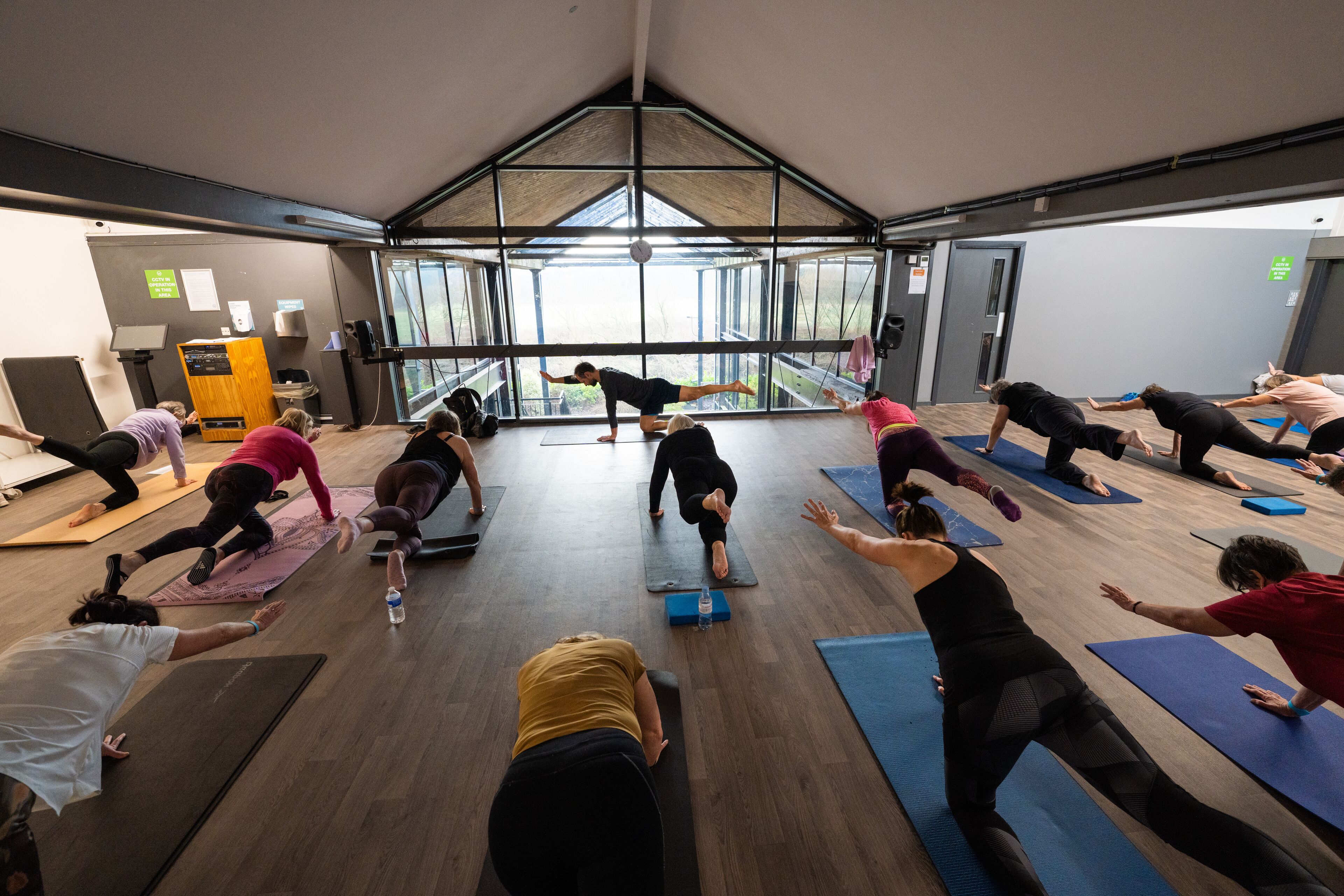 Un grupo de personas haciendo yoga en una habitación amplia con ventanas grandes y techo alto.