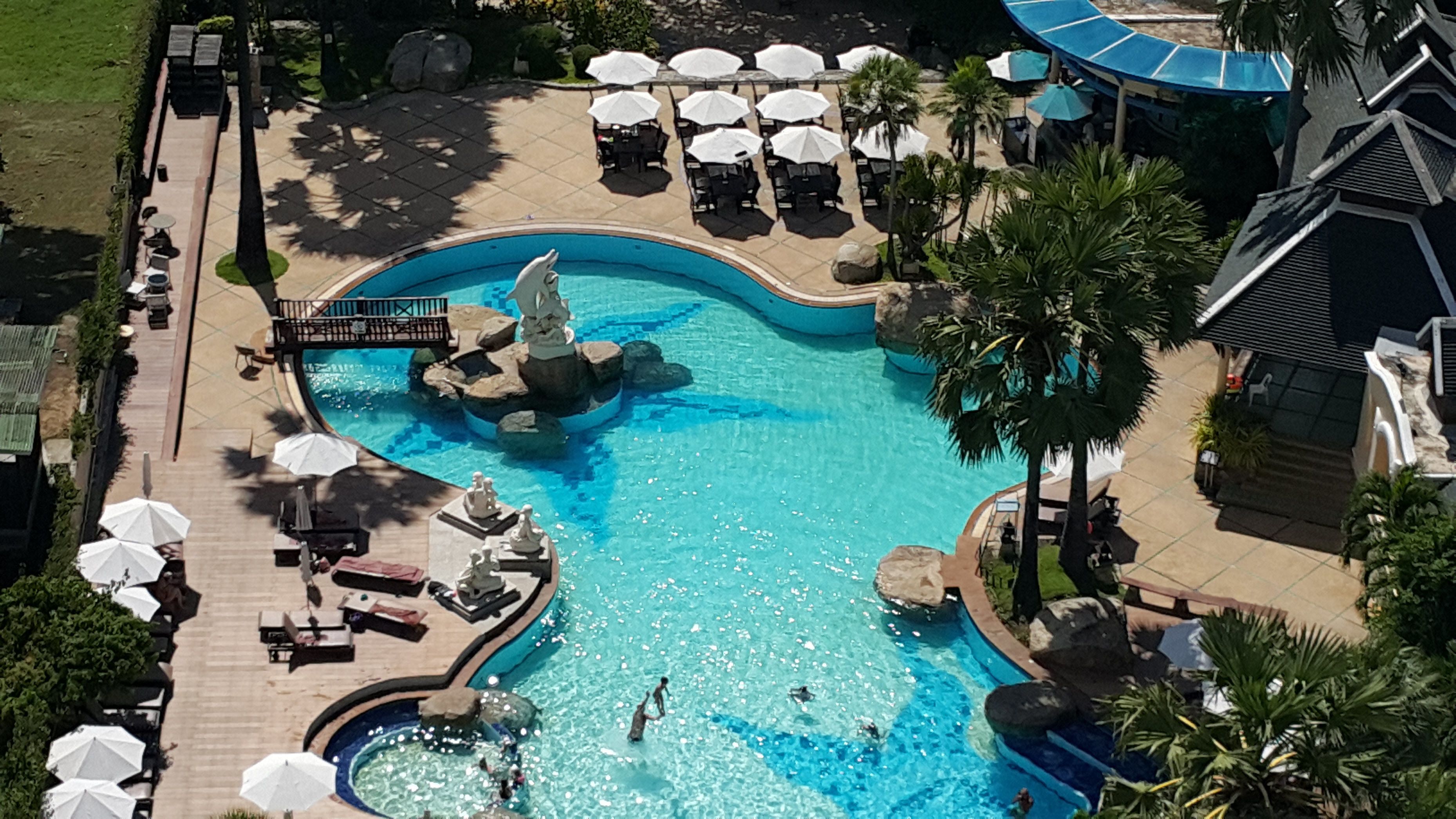 An aerial view of a resort with a large swimming pool, sun loungers, and palm trees.
