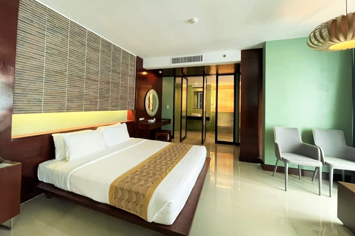 Habitación de hotel con una cama grande, un escritorio, una silla, un espejo y un baño visible a través de una puerta de cristal.
