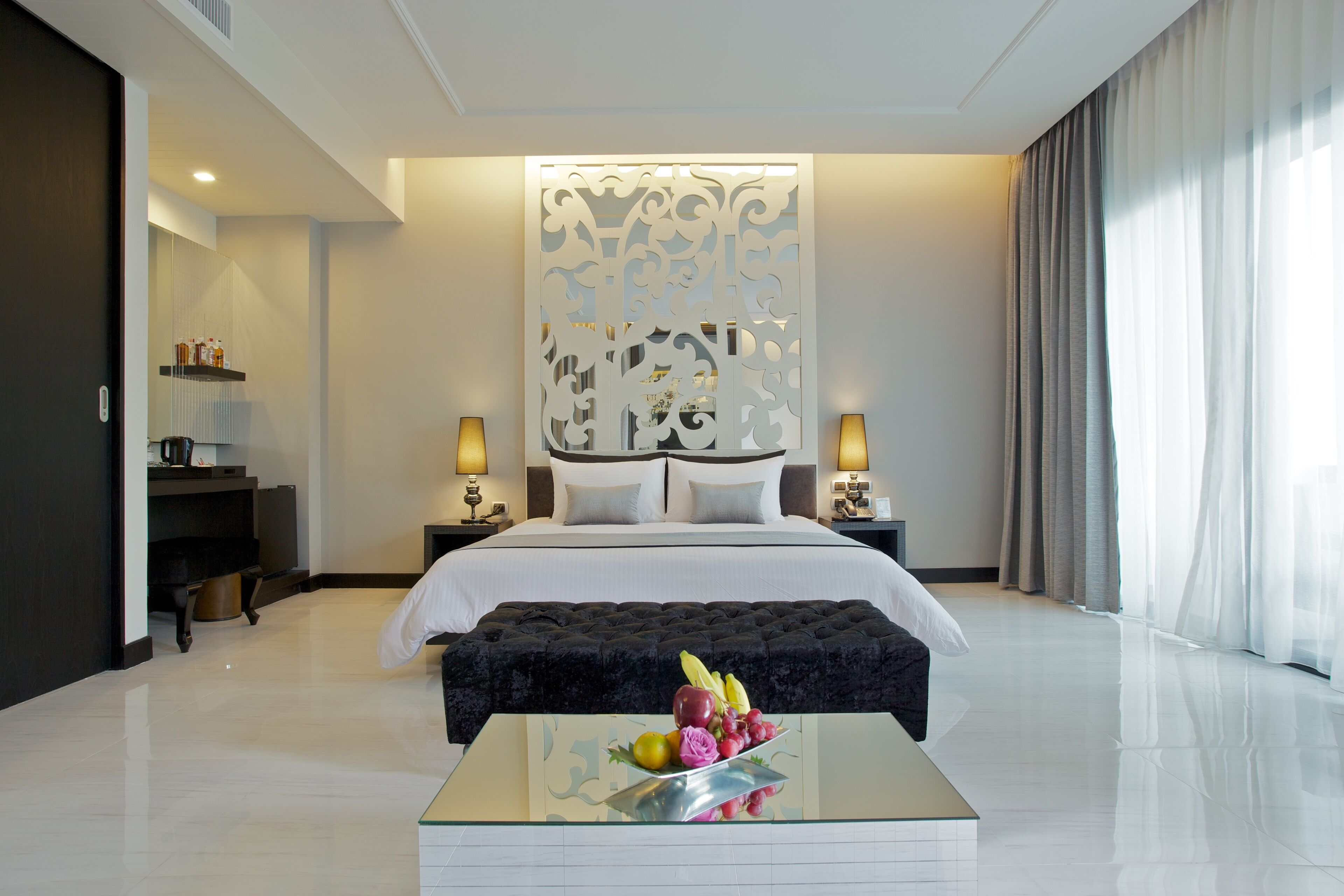 A modern hotel room with a large bed, a glass coffee table with fruit, a desk with a lamp, and a decorative wall panel.