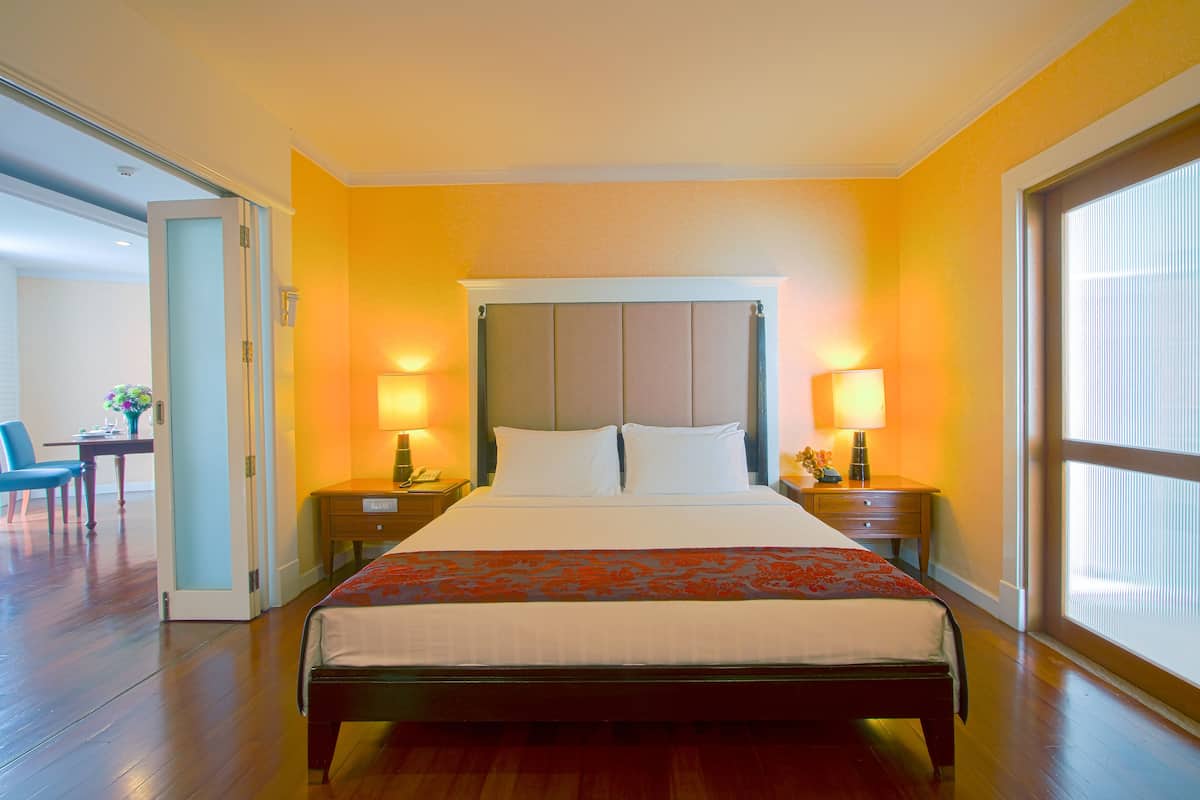 Habitación de hotel con una cama grande, dos mesitas de noche con lámparas, una mesita auxiliar y vistas al exterior a través de una ventana.