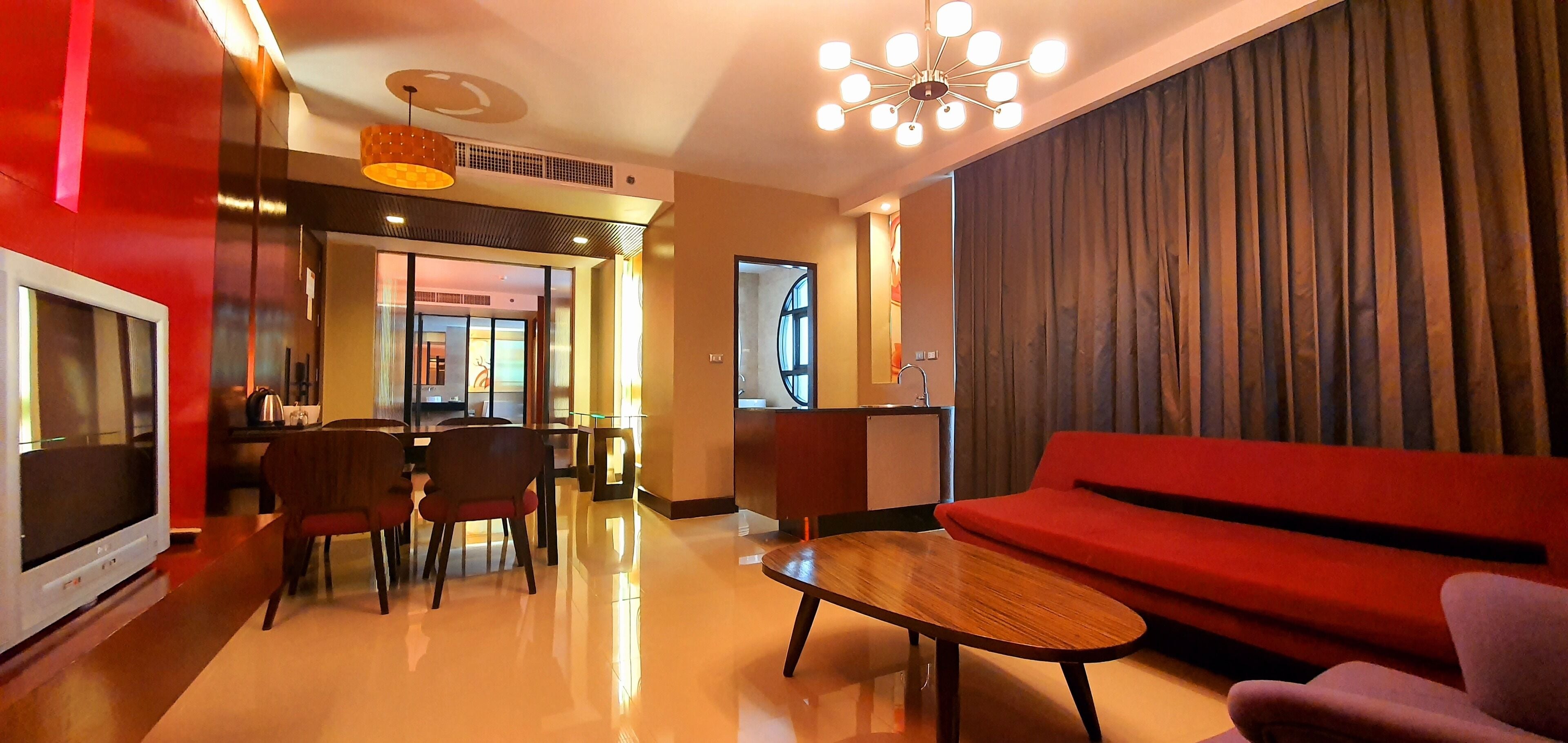 Un rebedor modern d'hotel amb un sofà vermell, una taula de centre de fusta i una televisió. Hi ha una zona de menjador amb caires i una taula, i una cuina al fons.