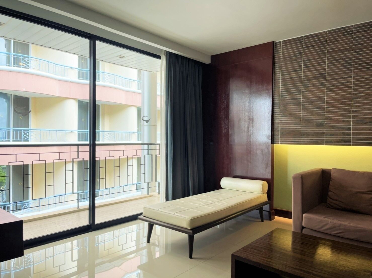 A modern hotel room with a large sliding glass door leading to a balcony, a sofa, a small table, and a wooden accent wall.