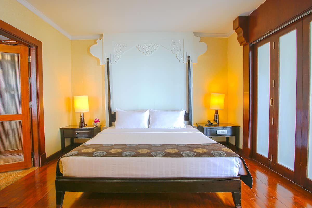 Una cama grande con cabecera, dos mesitas de noche con lámparas, piso de madera y una puerta corredera de cristal a la derecha.