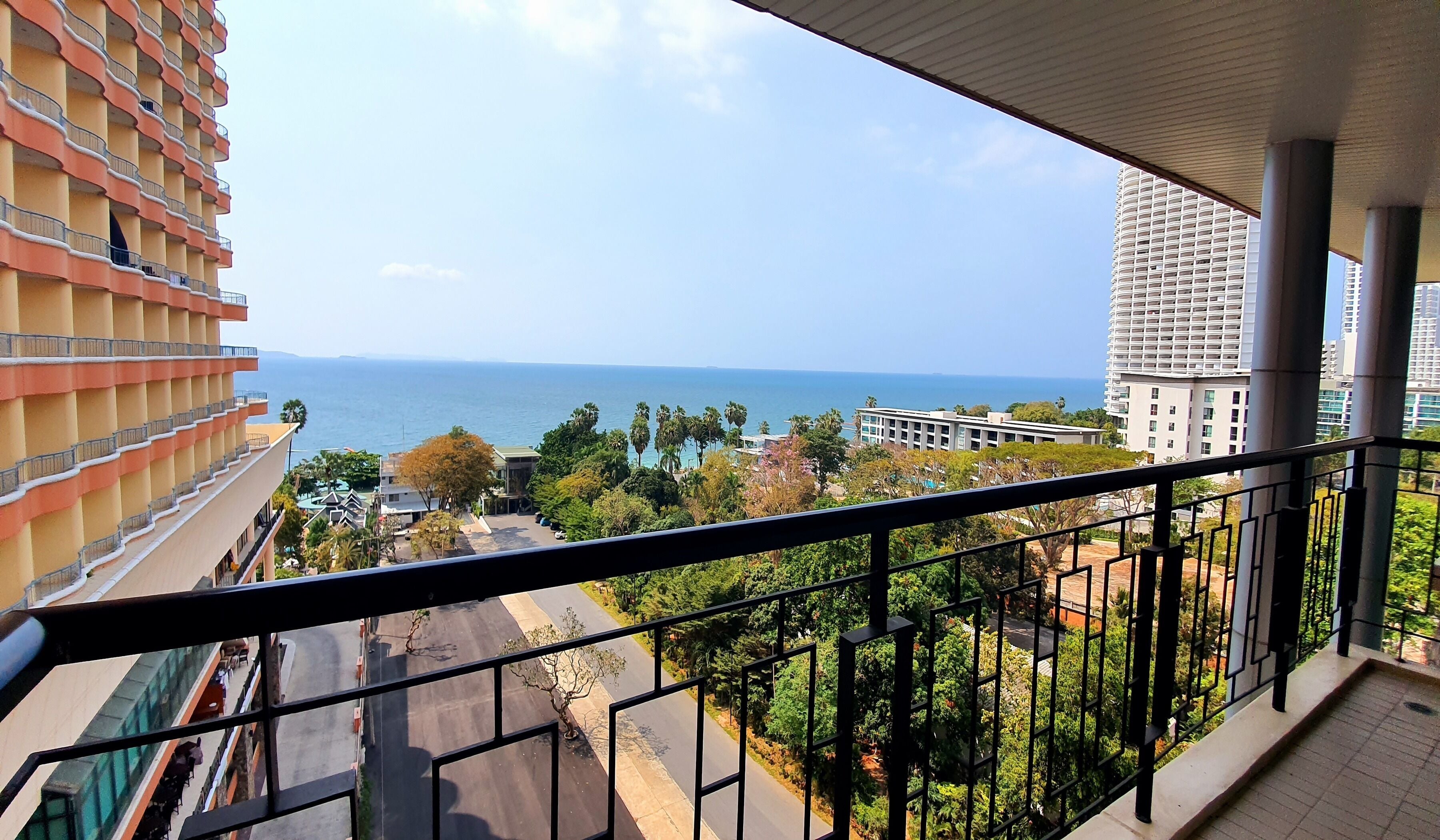 A balcony with a view of the sea, a building with balconies, and a road with greenery.