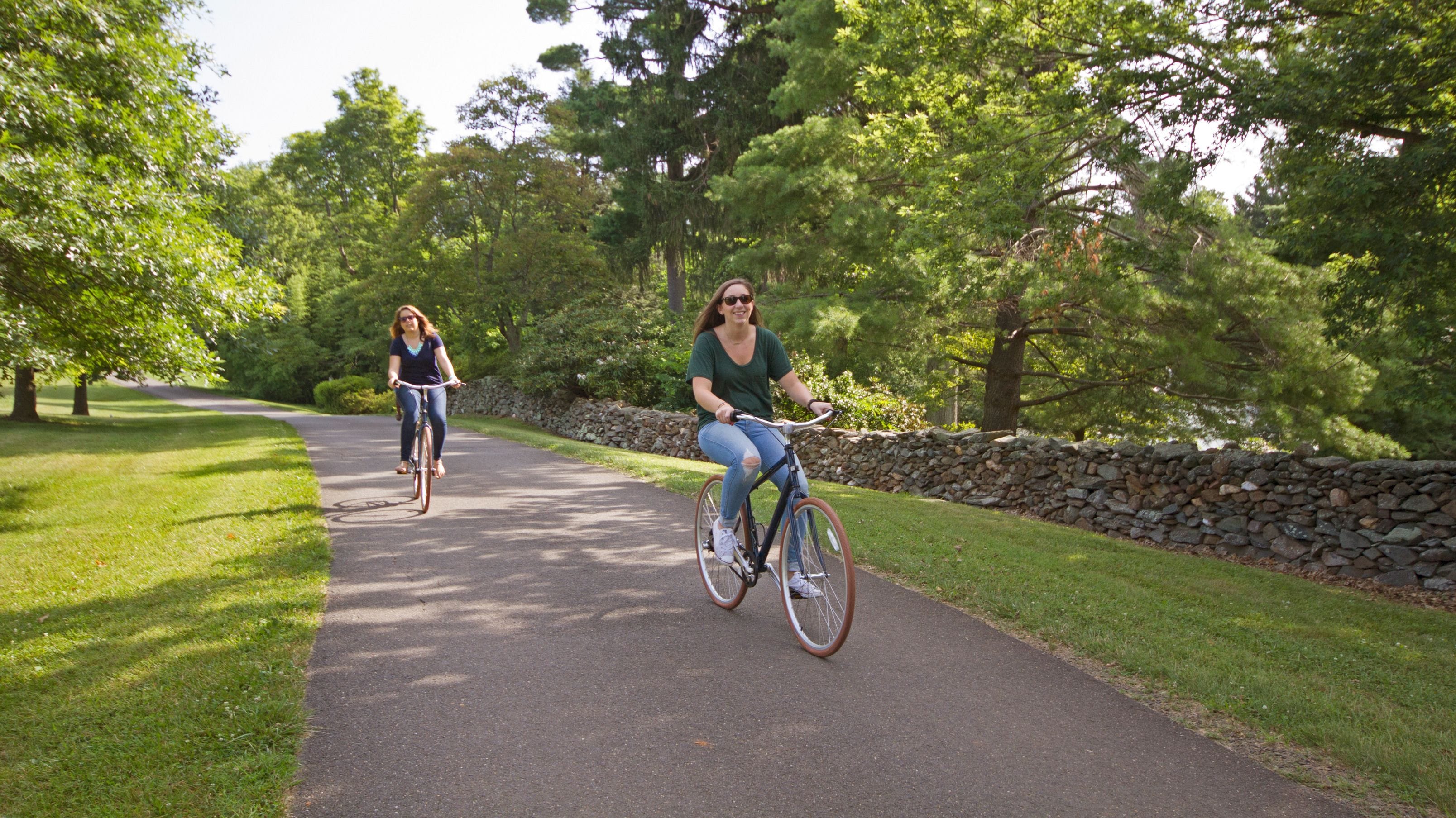 Two people riding bicycles on a paved path surrounded by greenery and stone walls.