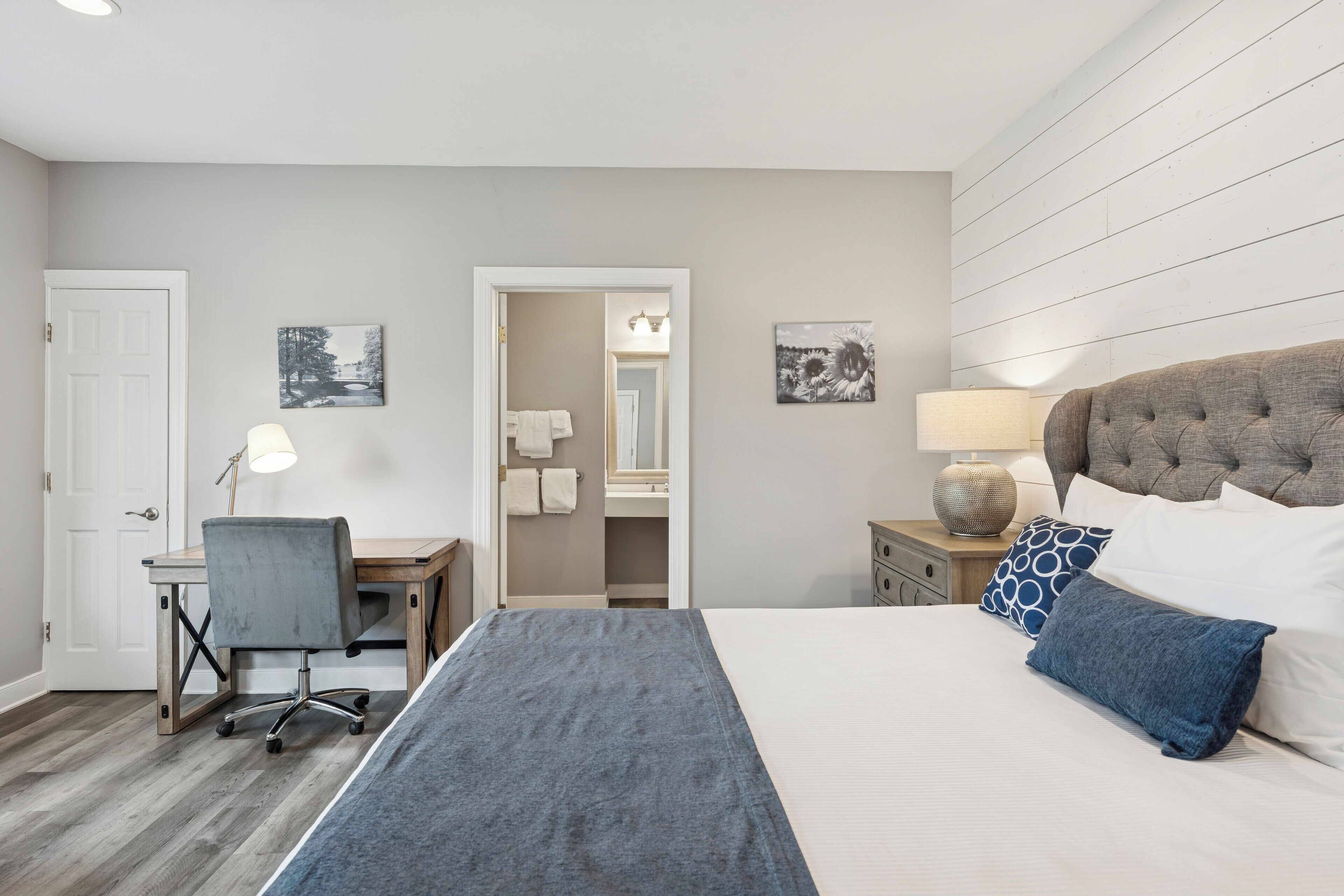 A bedroom with a large bed, a desk, a chair, and a nightstand with a lamp.