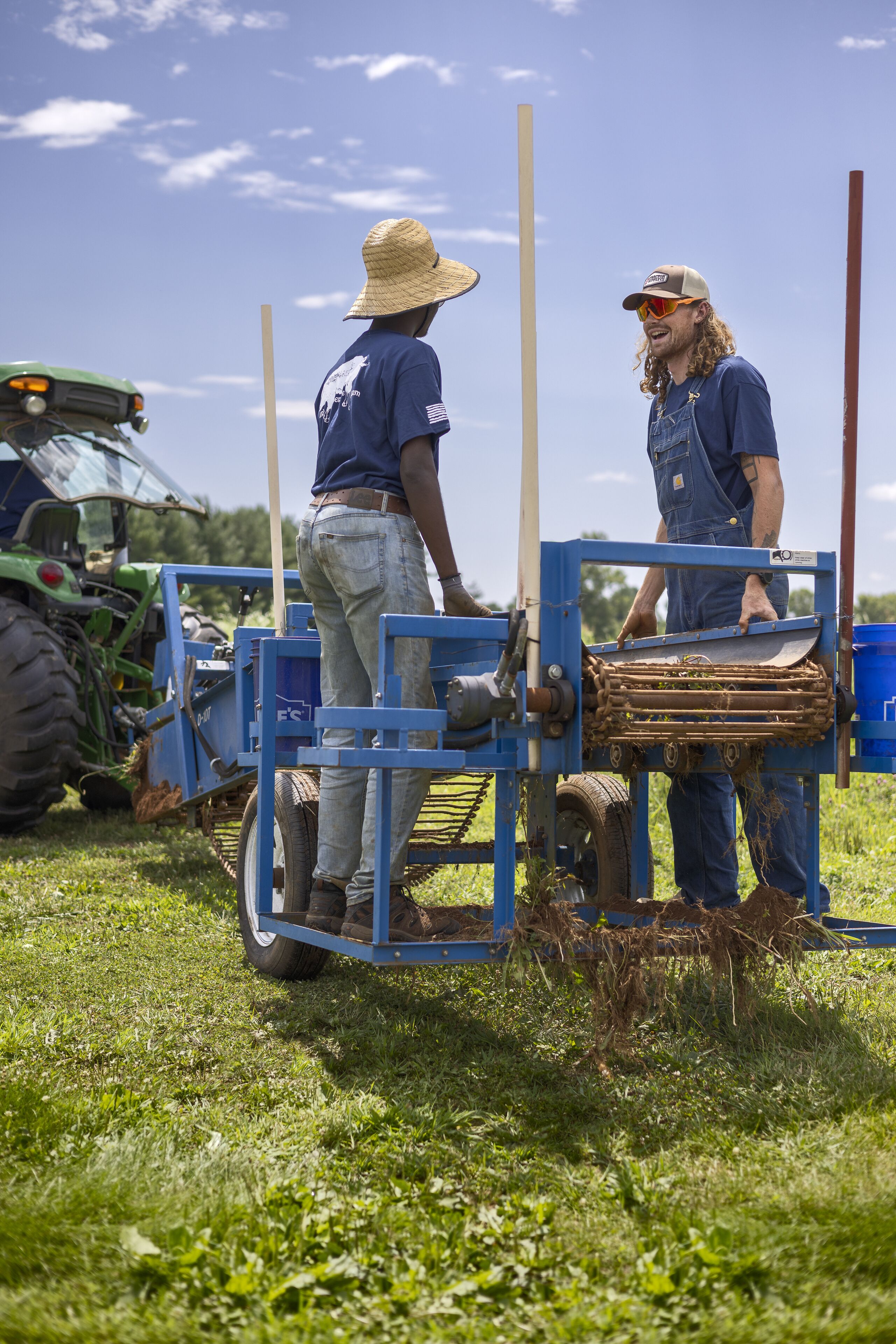 Two people standing next to a blue agricultural machine in a grassy field with a tractor in the background.