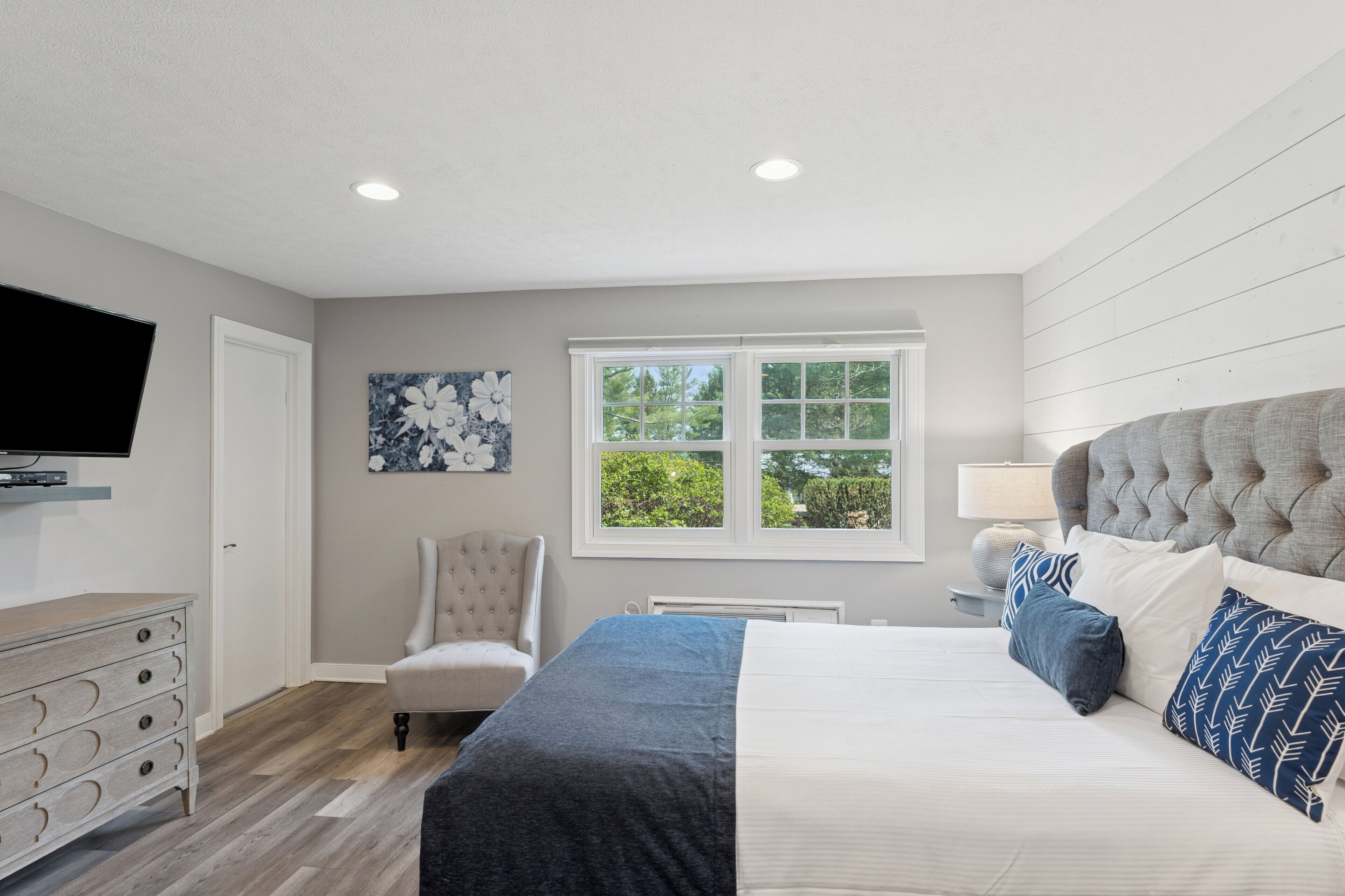 A bedroom with a bed, a chair, a dresser, a television, and a window with a view of trees.