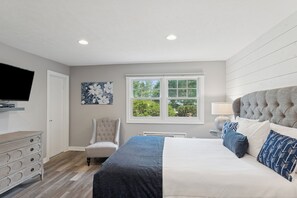 A bedroom with a bed, a chair, a dresser, a television, and a window with a view of trees. - Airlie Hotel (Warrenton)