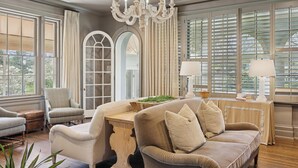 Un vaste salon comprenant un grand canapé beige, un fauteuil assorti et une table basse en bois. Un lustre est suspendu au plafond et la pièce est dotée de grandes fenêtres équipées de stores.