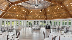Une grande salle avec de hauts plafonds, des poutres en bois et des guirlandes lumineuses. Des tables sont disposées, recouvertes de nappes blanches, et des chaises sont placées autour.