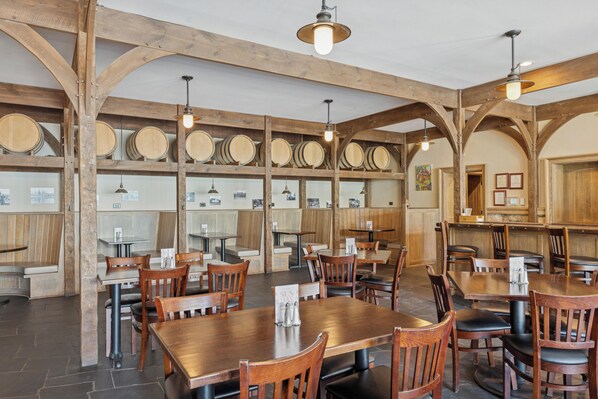 Un restaurante con vigas de madera, mesas y sillas. Se observan barriles de vino en estanterías y una barra con la carta.