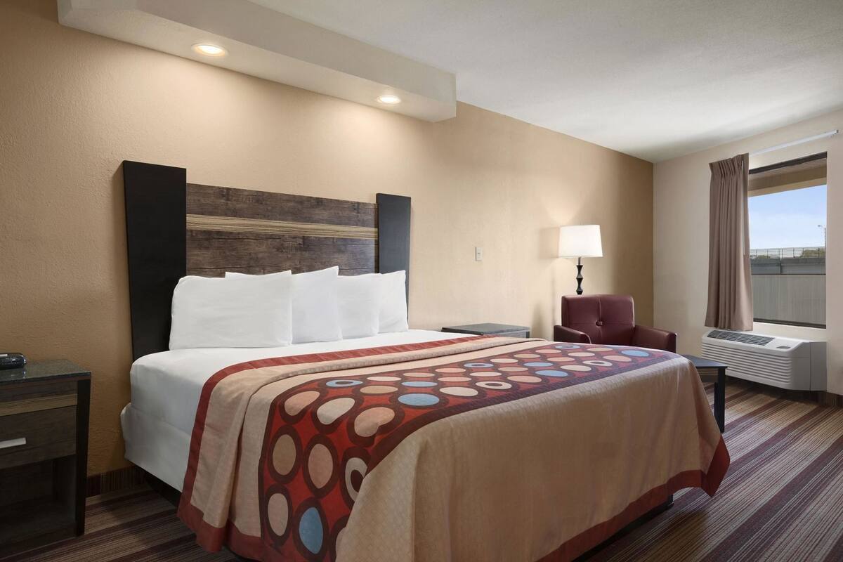 A hotel room with a bed, a desk, a chair, a lamp, and an air conditioning unit.