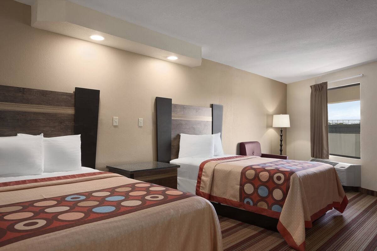 A hotel room with two beds, a desk, a chair, a window with curtains, and a lamp.