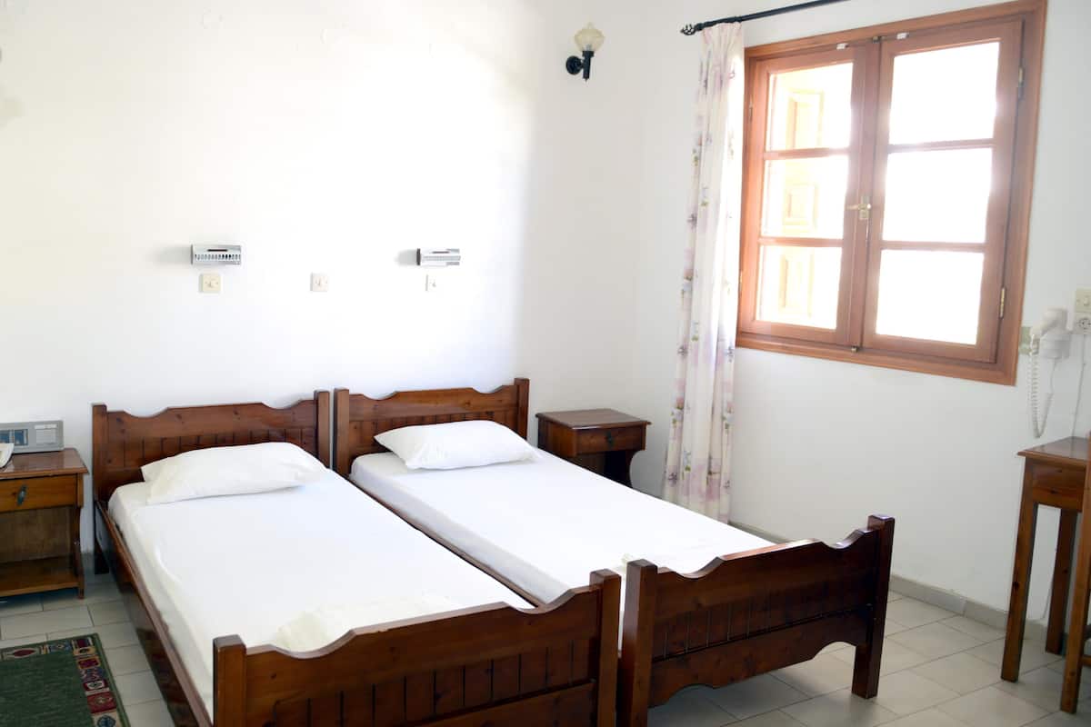 A bedroom with two wooden beds, a window with curtains, a bedside table, and a wall-mounted telephone.