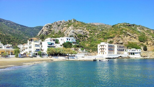 Une ville côtière avec des bâtiments qui bordent le front de mer, une mer d’un bleu limpide et un paysage montagneux en arrière-plan.
