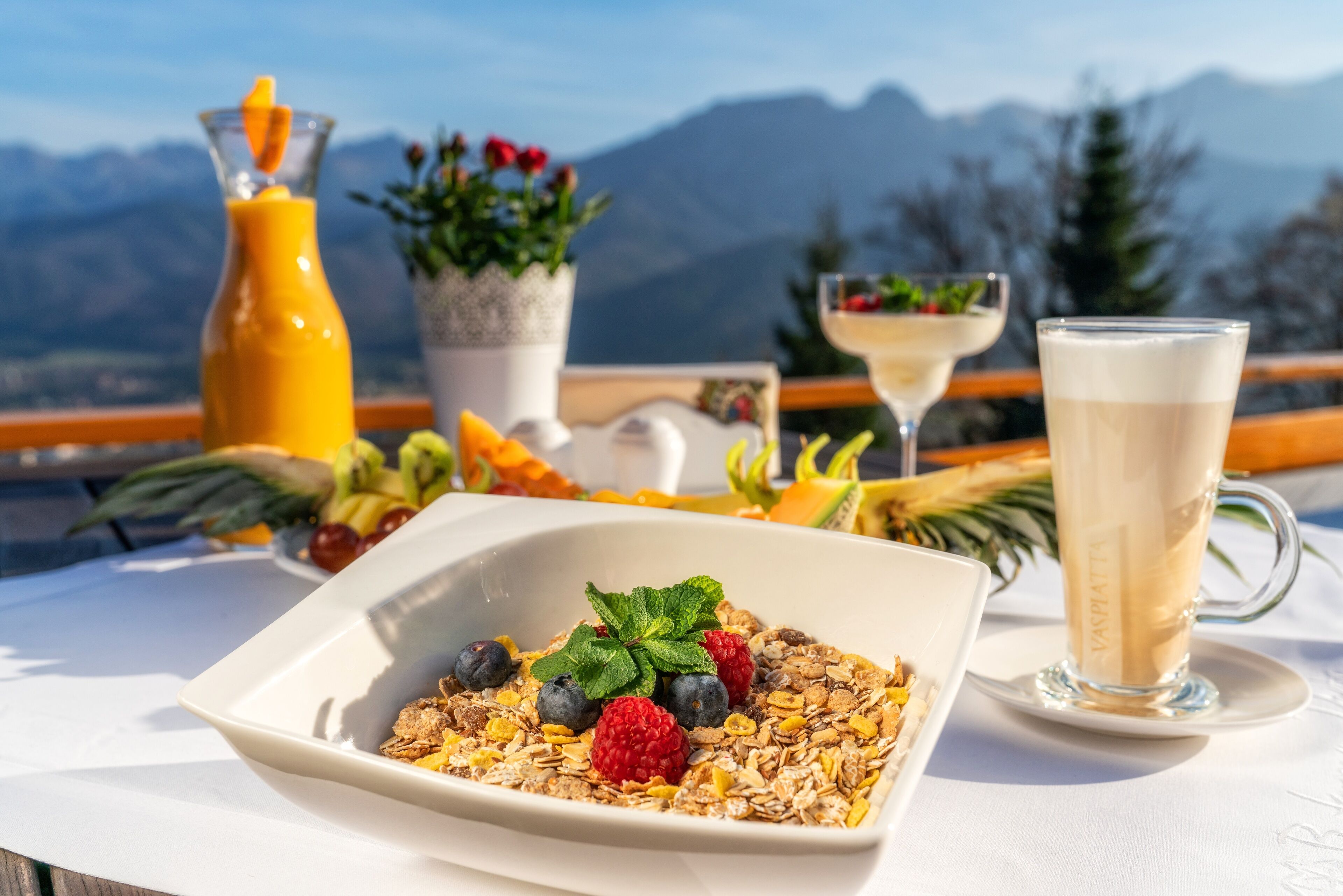 Ein Frühstückstisch mit einer Schüssel Müsli, einem Glas Milch und einem Fruchtsaft, mit Blick auf Berge im Hintergrund.