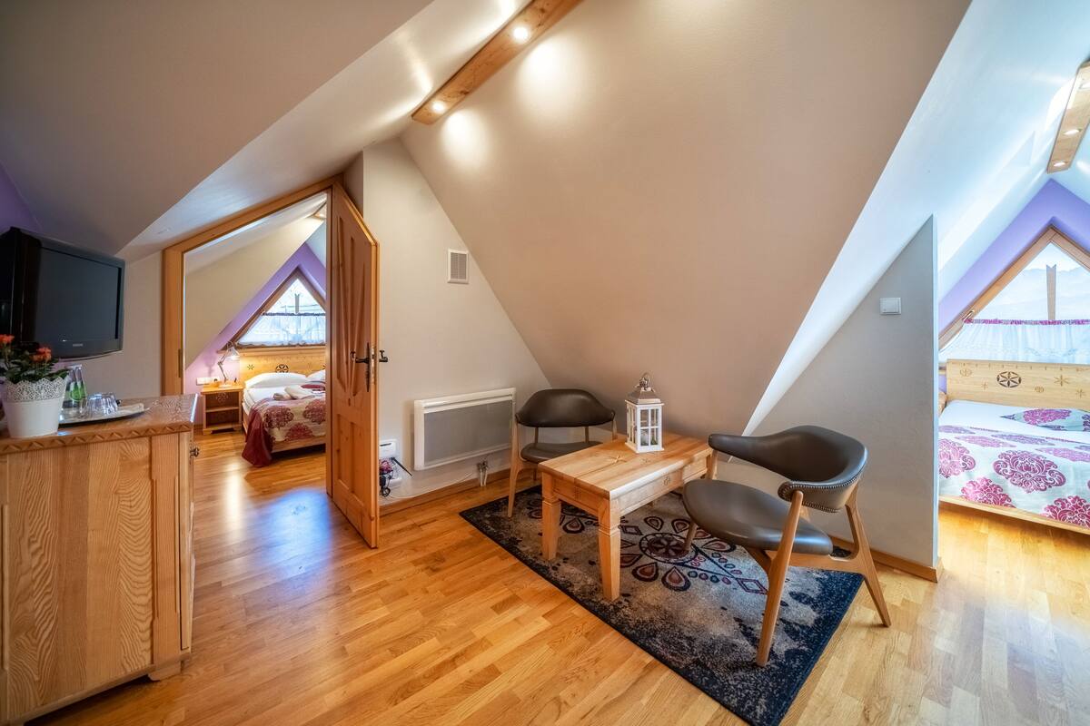 A cozy attic bedroom with a wooden desk, two chairs, a bed, and a TV.