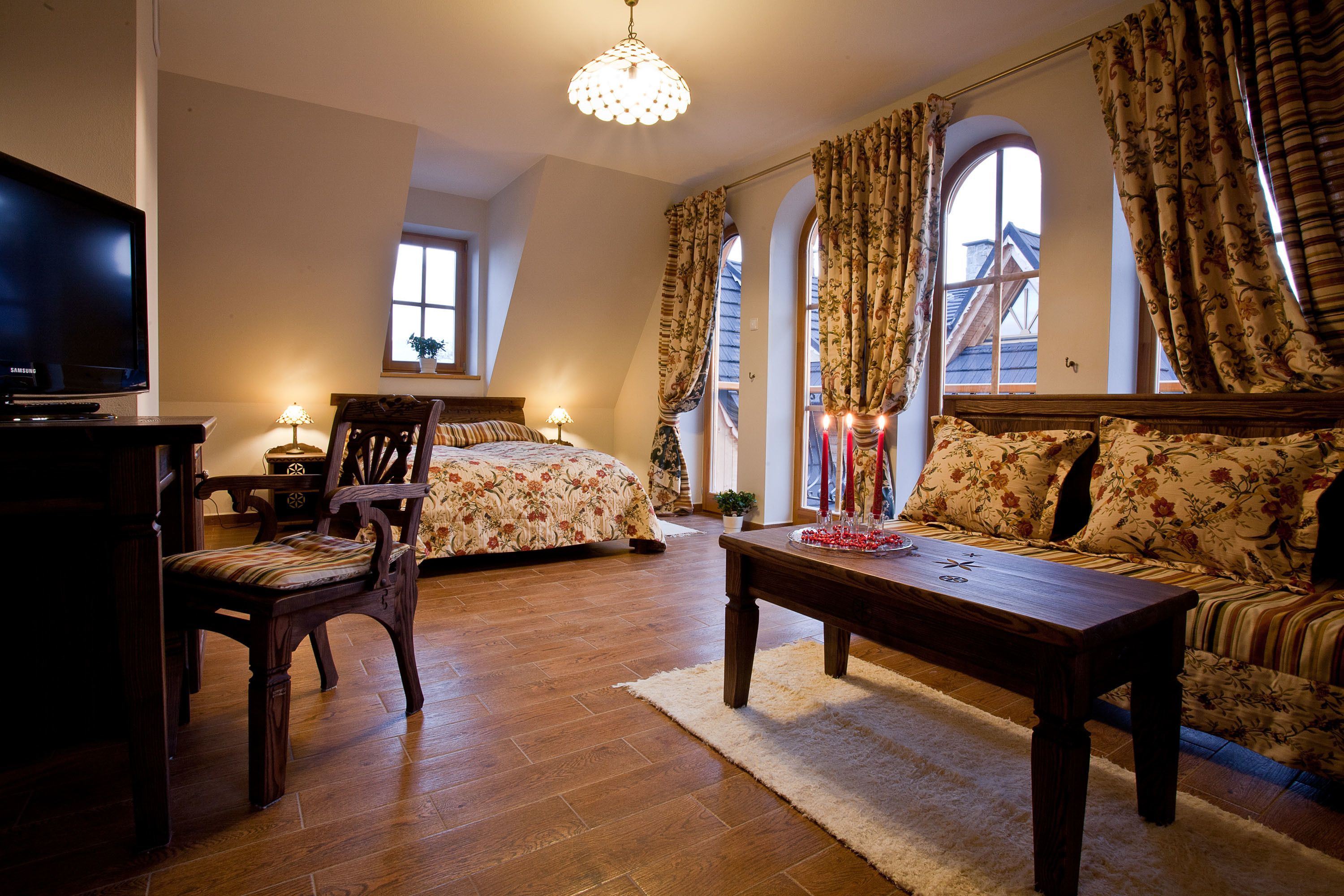 Ein gemütliches Zimmer mit Holzboden, einer Couch, einem Bett mit Blumenmuster und einem Fernseher, sowie einer Kristallleuchter.