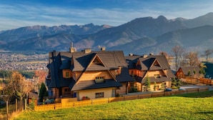 Una casa de estilo chalete con varios techos a dos aguas, rodeada por una valla de madera, con montañas y un valle de fondo.
