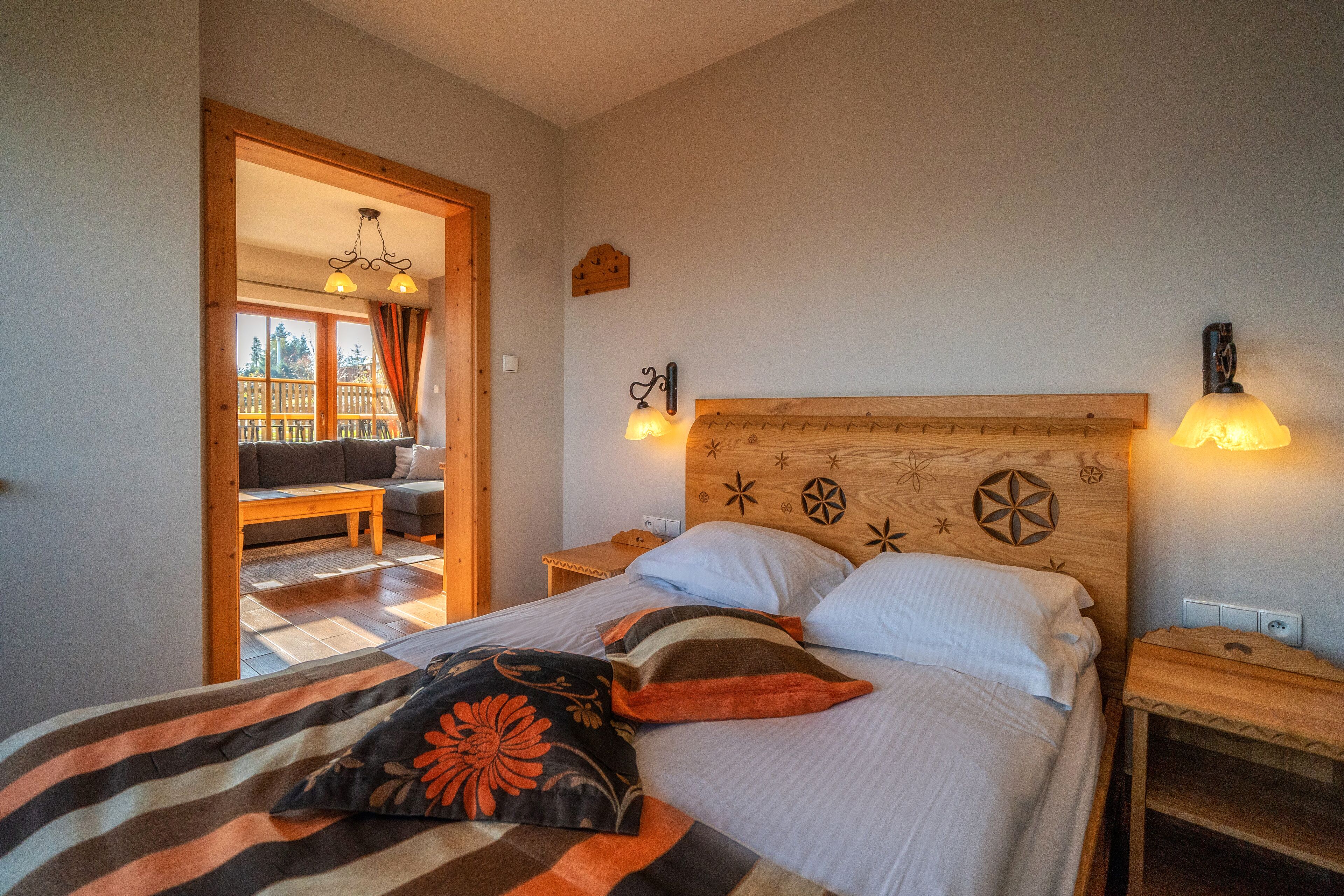 A bedroom with a wooden headboard, two bedside lamps, a bed with a striped blanket and pillows, a nightstand, and a view of a balcony with a wooden fence and a chair.