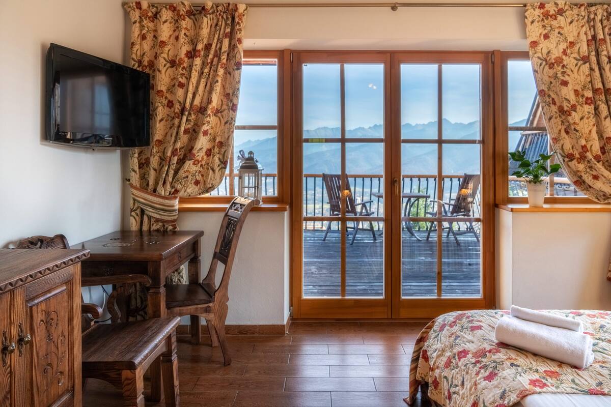 A room with a wooden desk, a bed with a floral bedspread, and a balcony with a view of mountains.