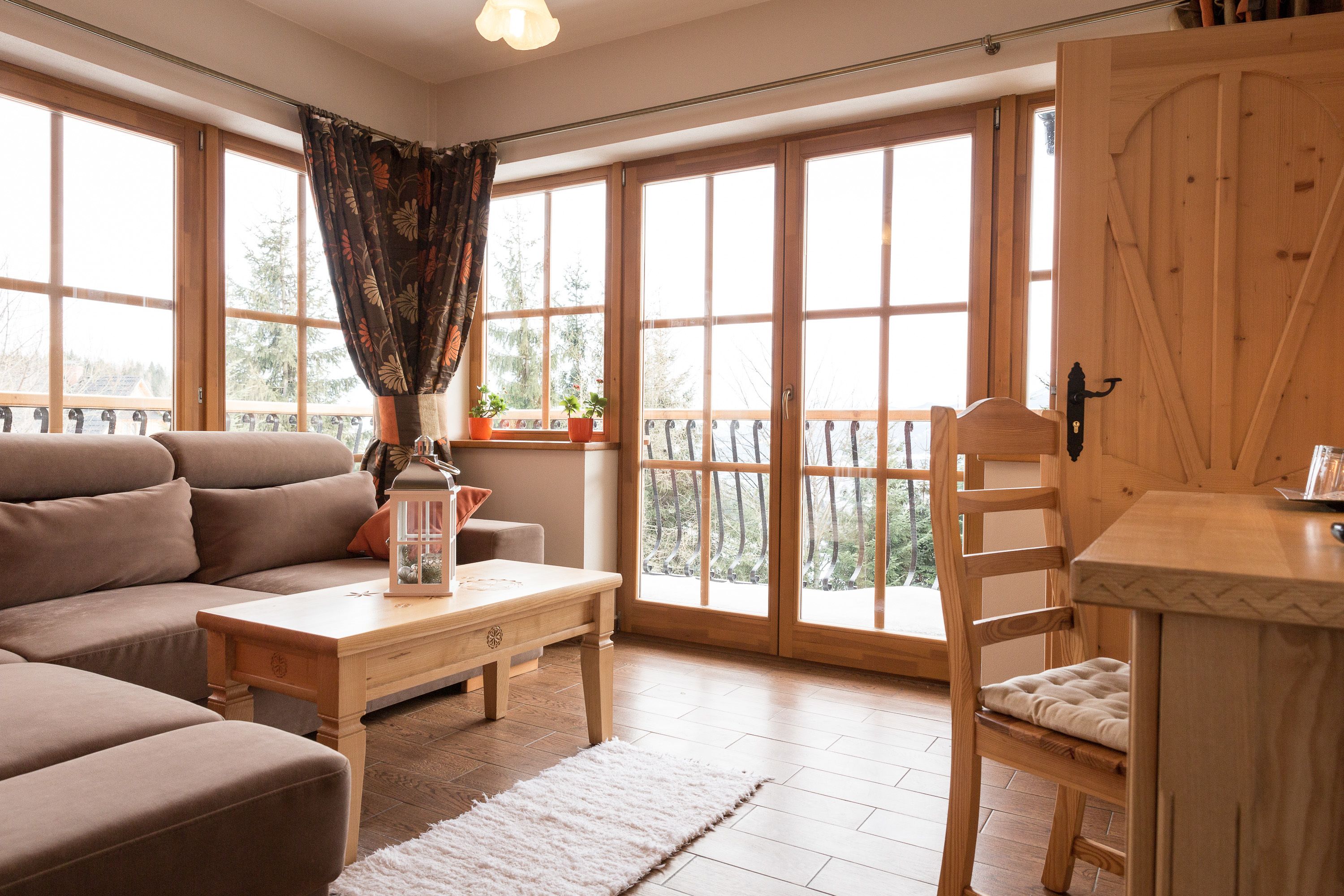 Un salon chaleureux avec un canapé, une table en bois et une chaise. De grandes fenêtres sont ornées de rideaux, et il y a un balcon avec vue sur les arbres.