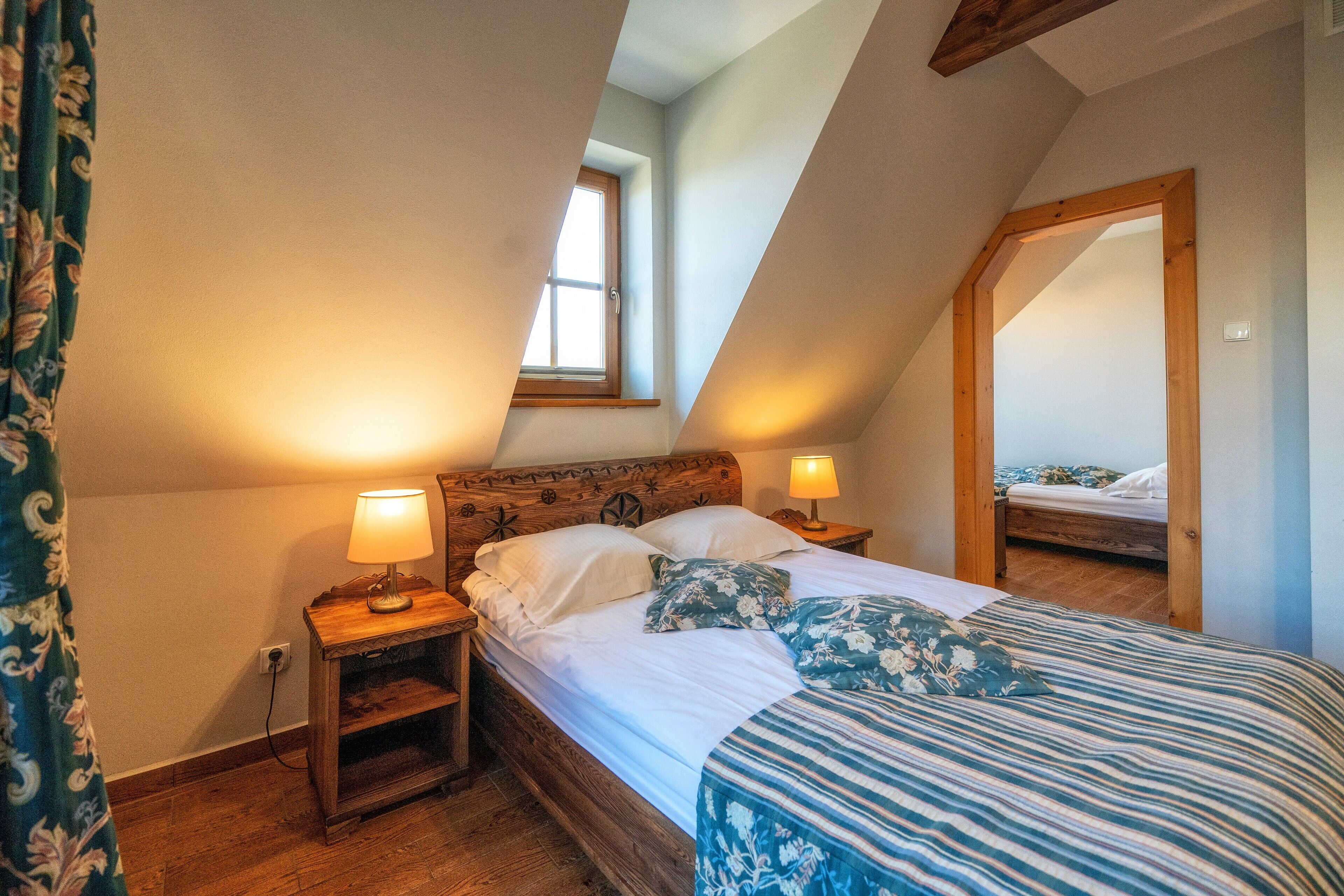 A bedroom with a sloped ceiling, wooden headboard, two bedside lamps, a bed with white bedding and floral pillows, a window, and a mirror on the wall.