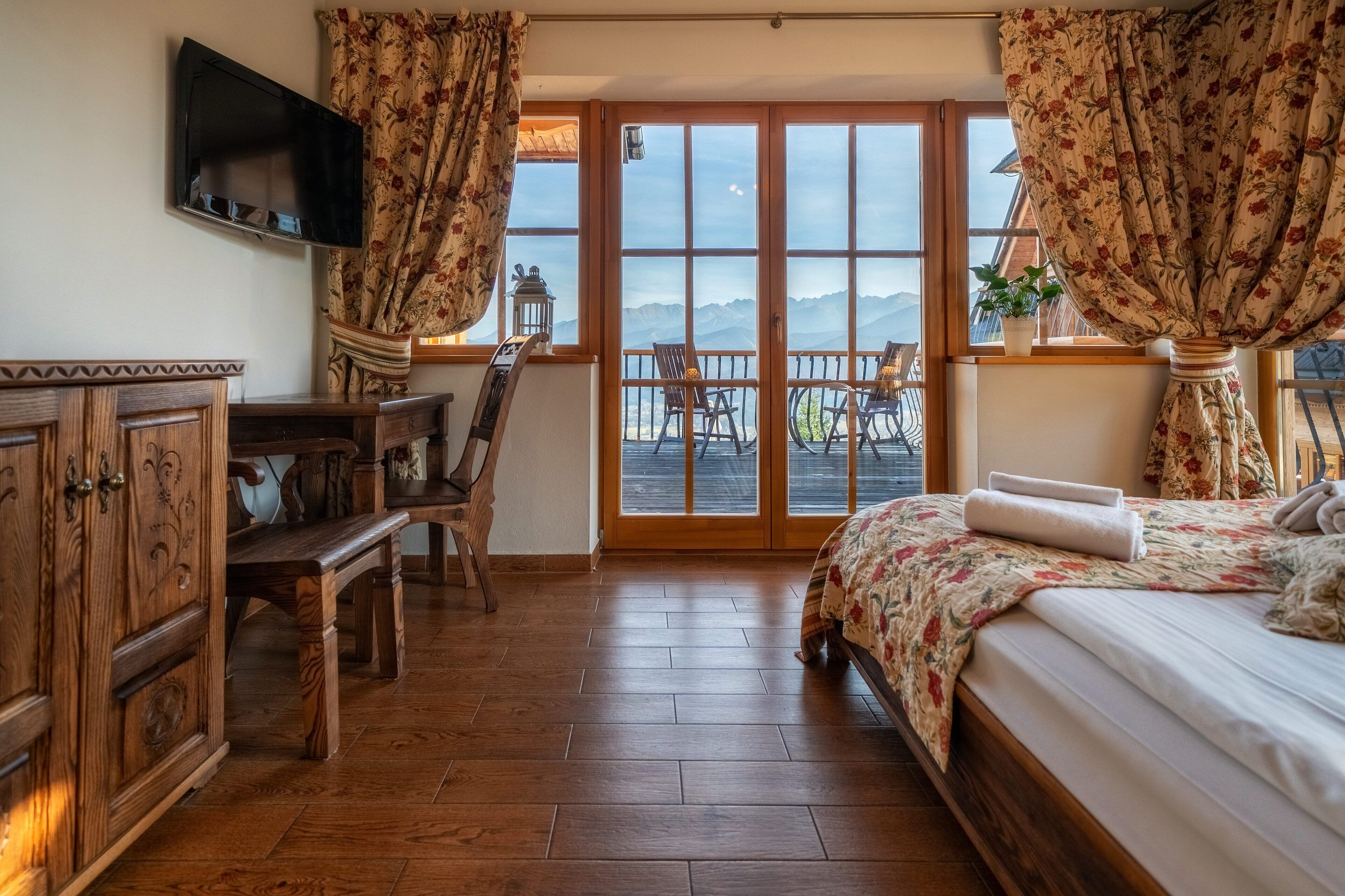 Una camera da letto con pavimento in legno, un letto con copripiumino floreale, una scrivania con sedia, un televisore a parete e un balcone con sedie e una pianta in vaso.