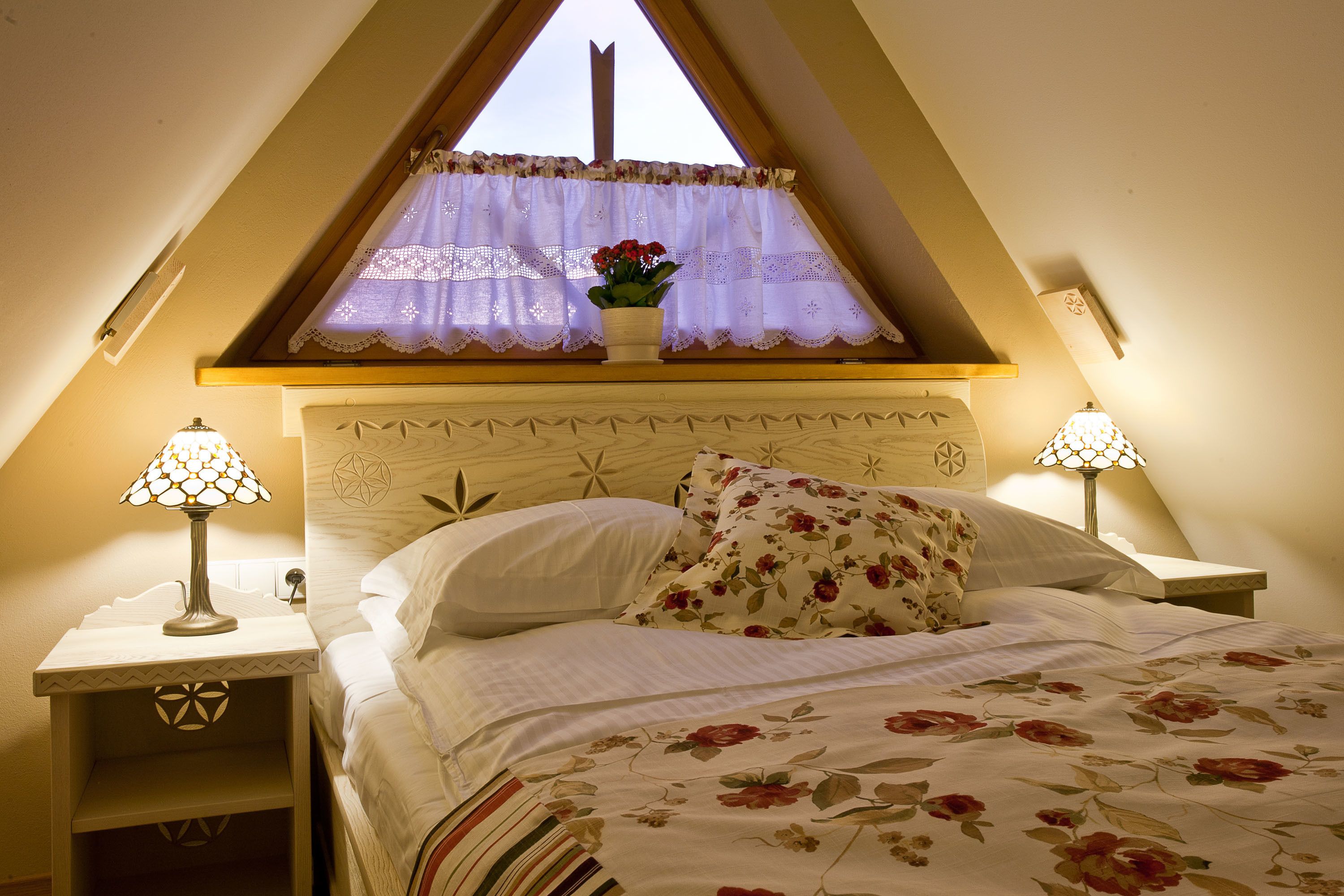 A bedroom with a bed, two bedside lamps, a window with lace curtains, and a floral bedspread.