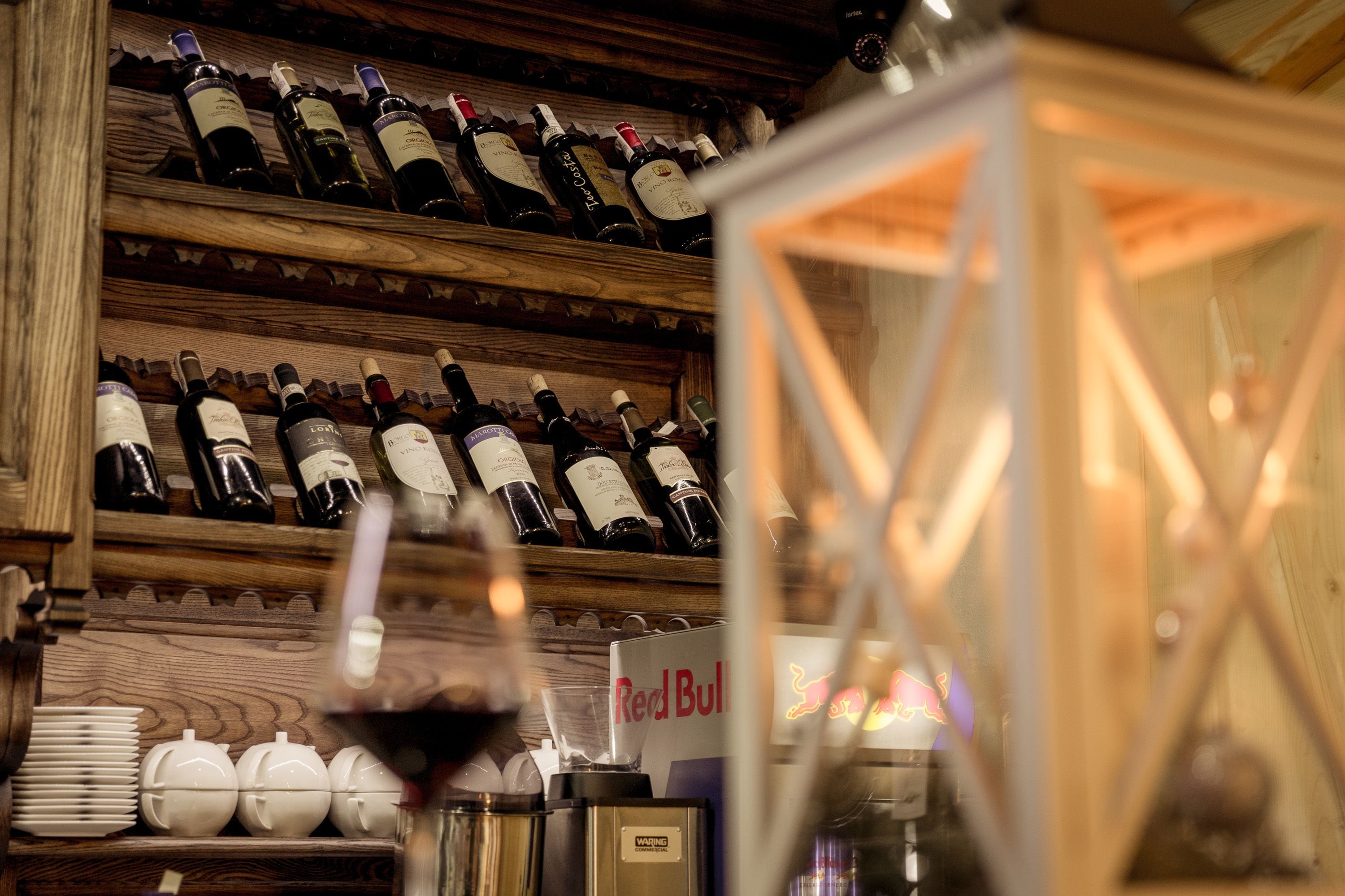 A wine rack with multiple bottles, a Red Bull refrigerator, and a glass of red wine.