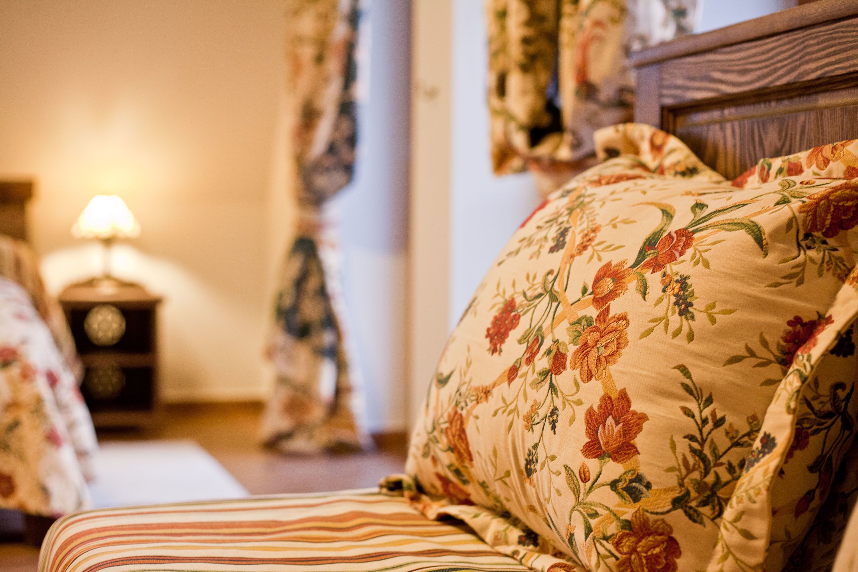 A bedroom with a floral-patterned bedspread, a striped pillow, and a nightstand with a lamp.