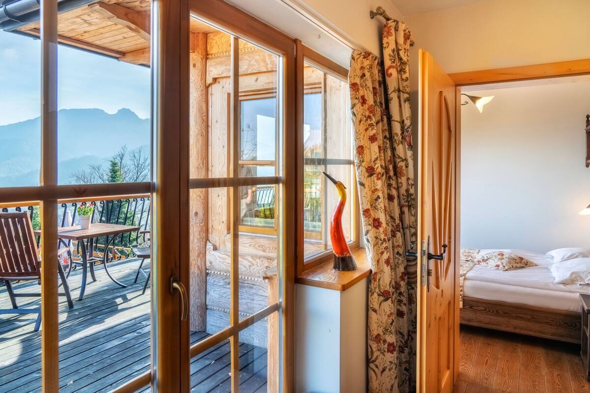 A wooden deck with a table and chairs, a bed with a floral bedspread, and a view of mountains through large windows.