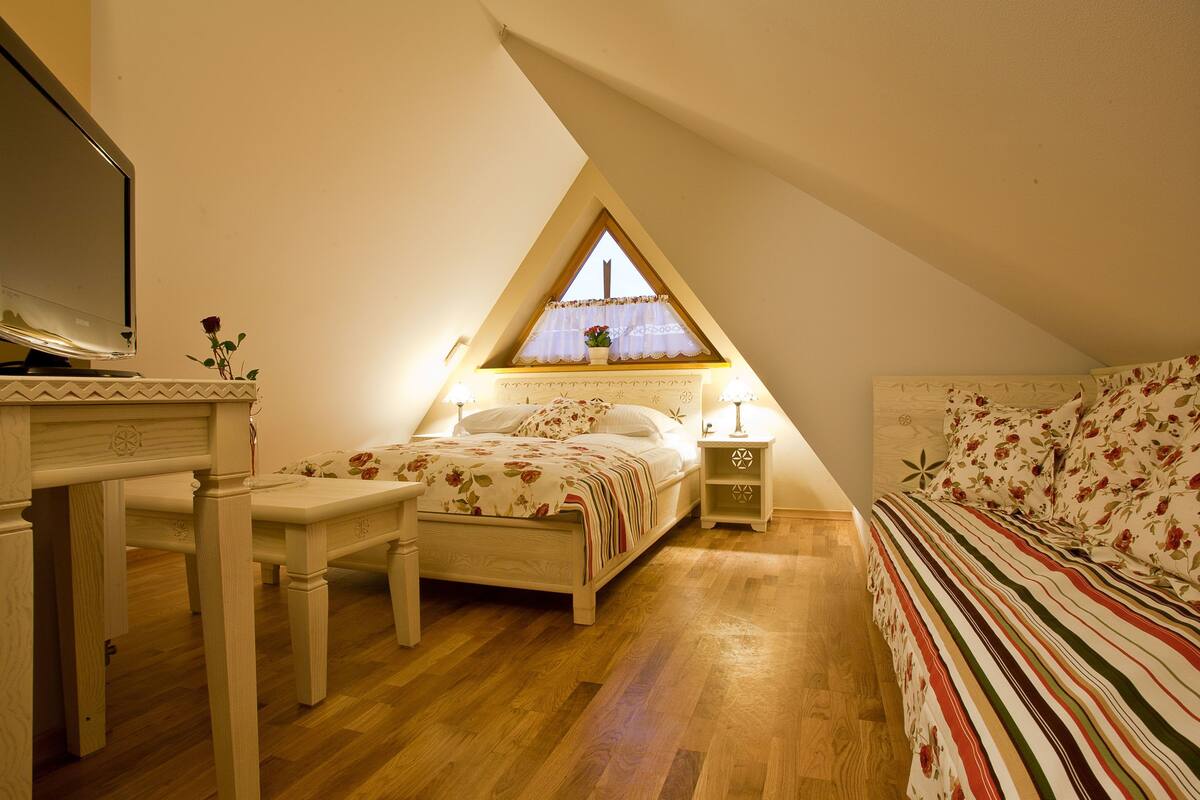 A bedroom with a wooden floor, a bed with floral bedding, a nightstand, a dresser, and a window with a triangular ceiling.