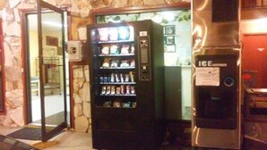 Una máquina expendedora con variedad de aperitivos y bebidas, un refrigerador con dispensador de hielo, y una pared de piedra con puerta de cristal.