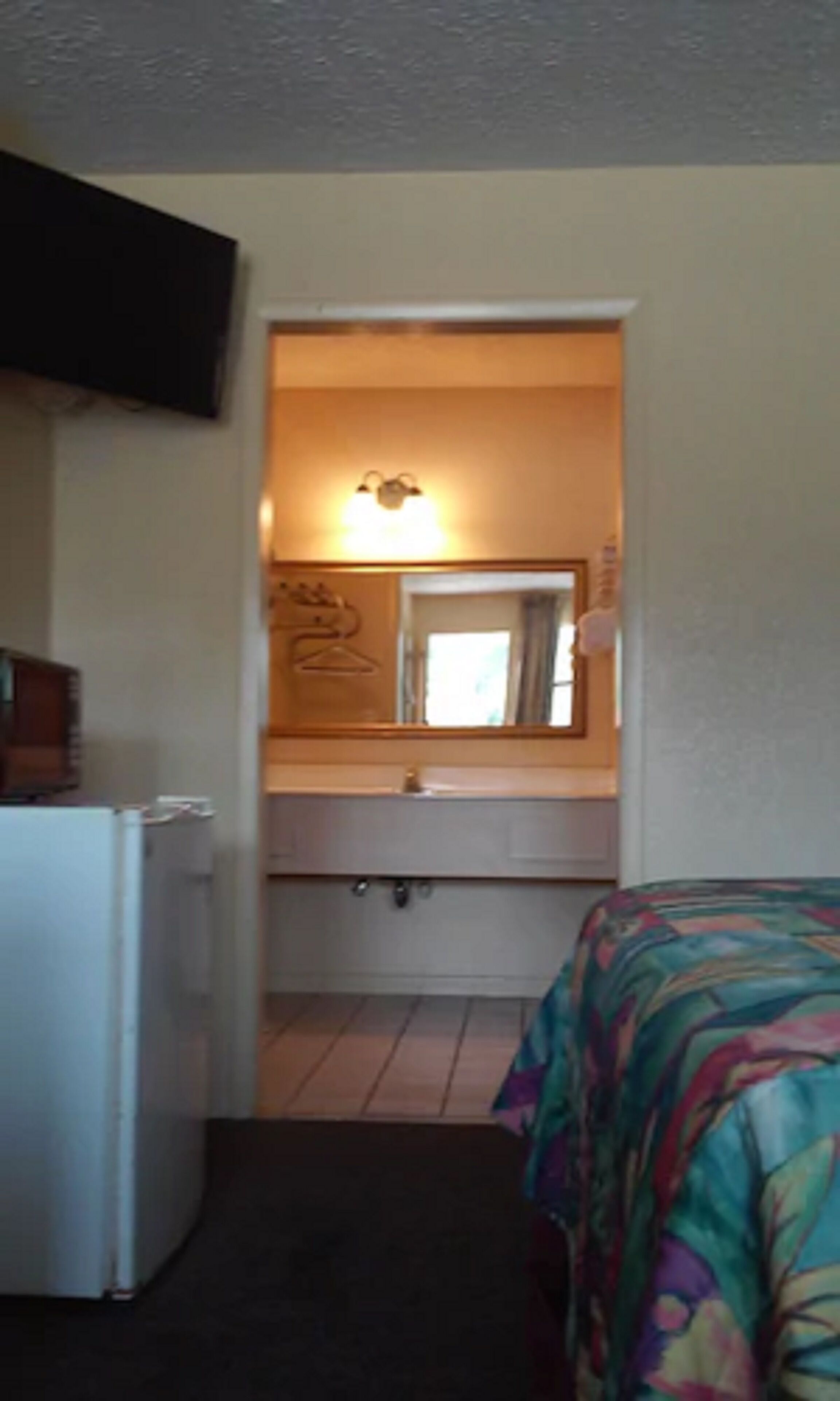 A compact hotel room with a bed, a small kitchenette, a mirror, and a television mounted on the wall.
