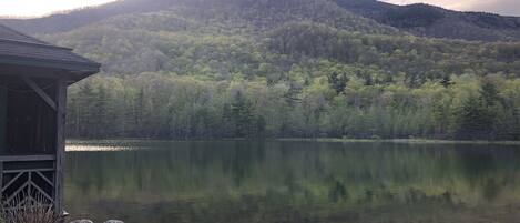 A cabin with a stone deck and a view of a lake and forested mountains.