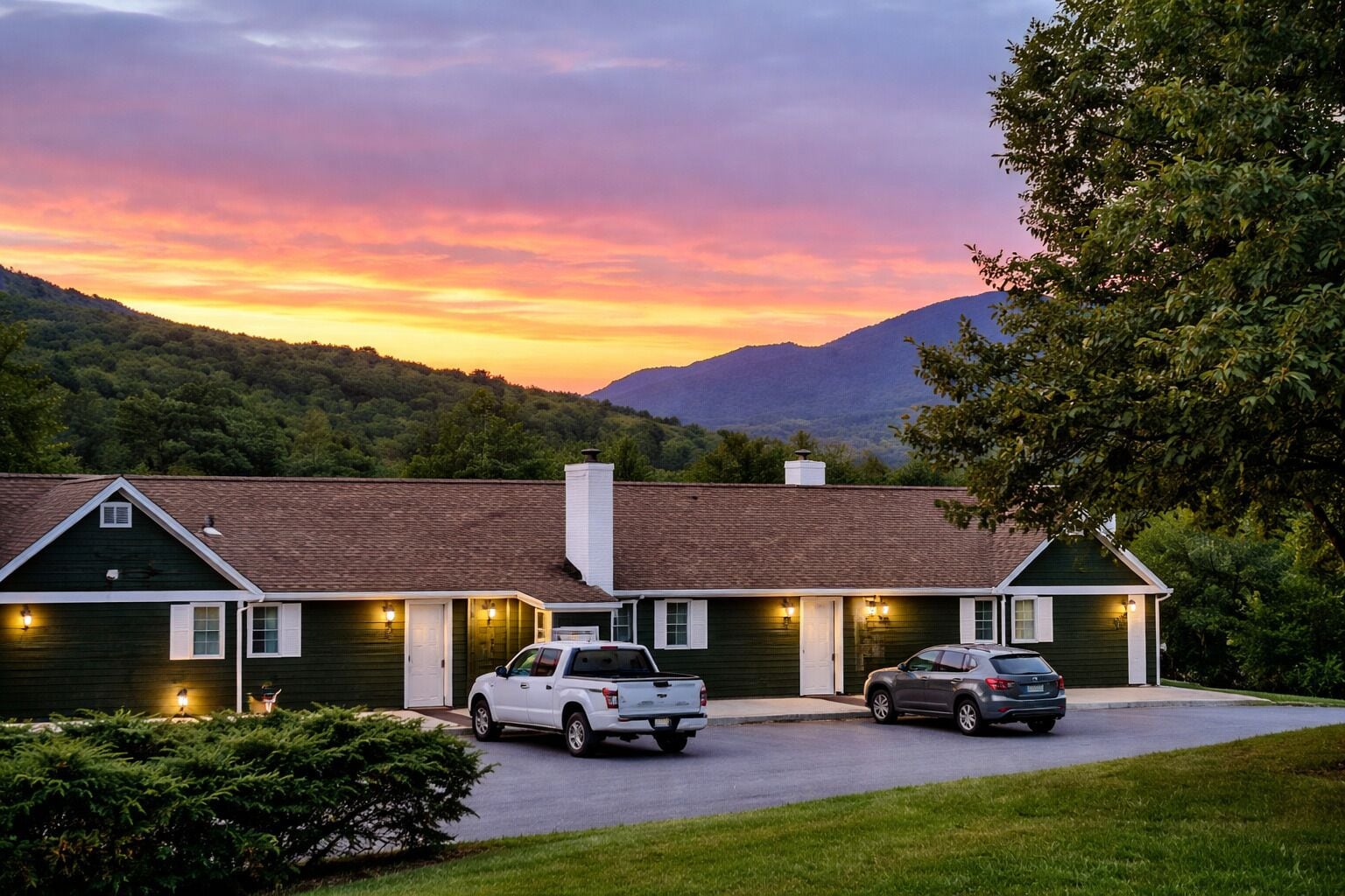 A two-story building with a green exterior, a driveway with two parked vehicles, and a mountainous backdrop during sunset.