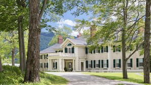 Une grande maison à deux étages, dotée d’une entrée centrale et entourée d’arbres, avec une chaîne de montagnes en toile de fond.