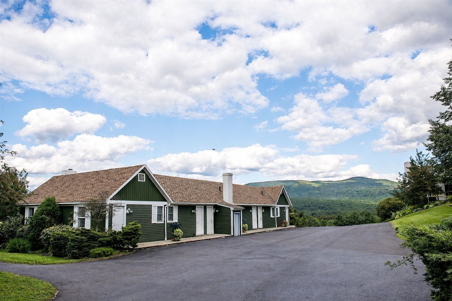 A two-story building with a green facade and a brown roof, a driveway leading to the entrance, and a mountainous backdrop.
