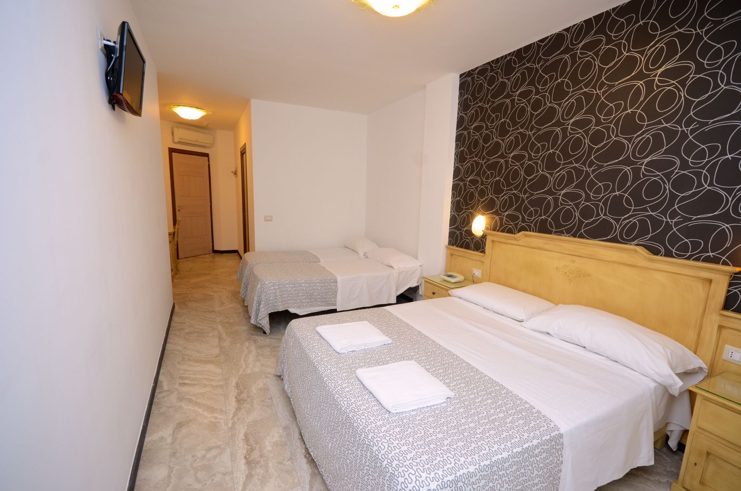 A hotel room with two beds, a wooden headboard, a patterned wall, and a TV mounted on the wall.